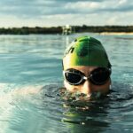 A determined swimmer in goggles and cap emerges from a tranquil lake on a sunny day.