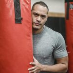 Adult serious ethnic male athlete with muscles in t shirt looking at camera between punching bags in gymnasium